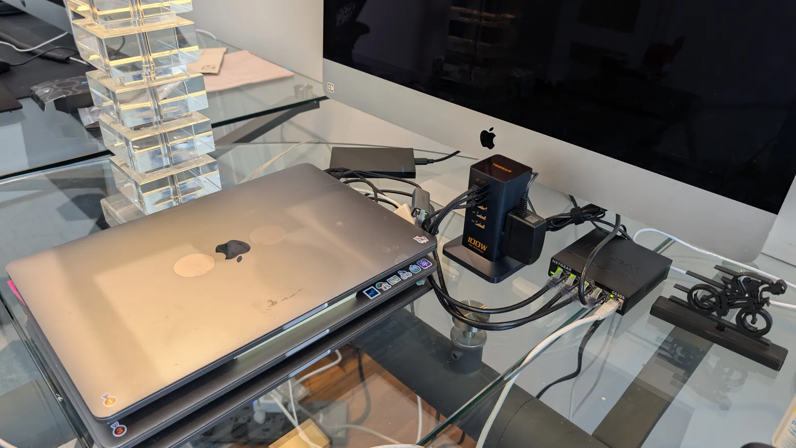 A well-used MacBook connected to an iMac, network equipment, and power adapters on a glass desk.